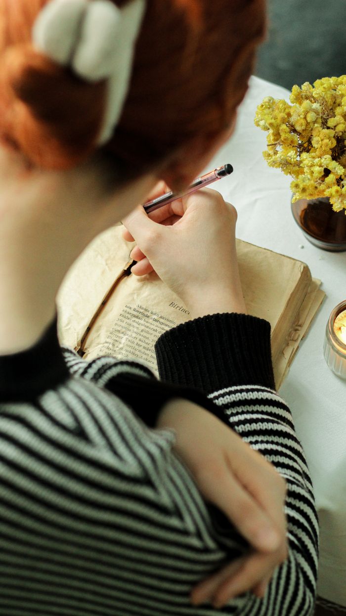 A woman with red hair writes in an old book while seated at a table with flowers and a candle.